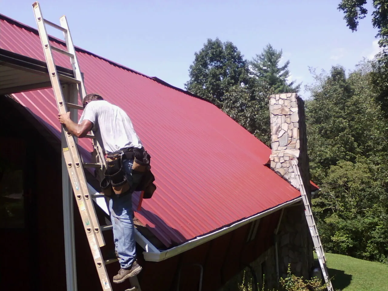Roofer on a ladder beside a red metal roof and stone chimney — Soffit & Fascia Repair by Roofing Brothers in Seaside